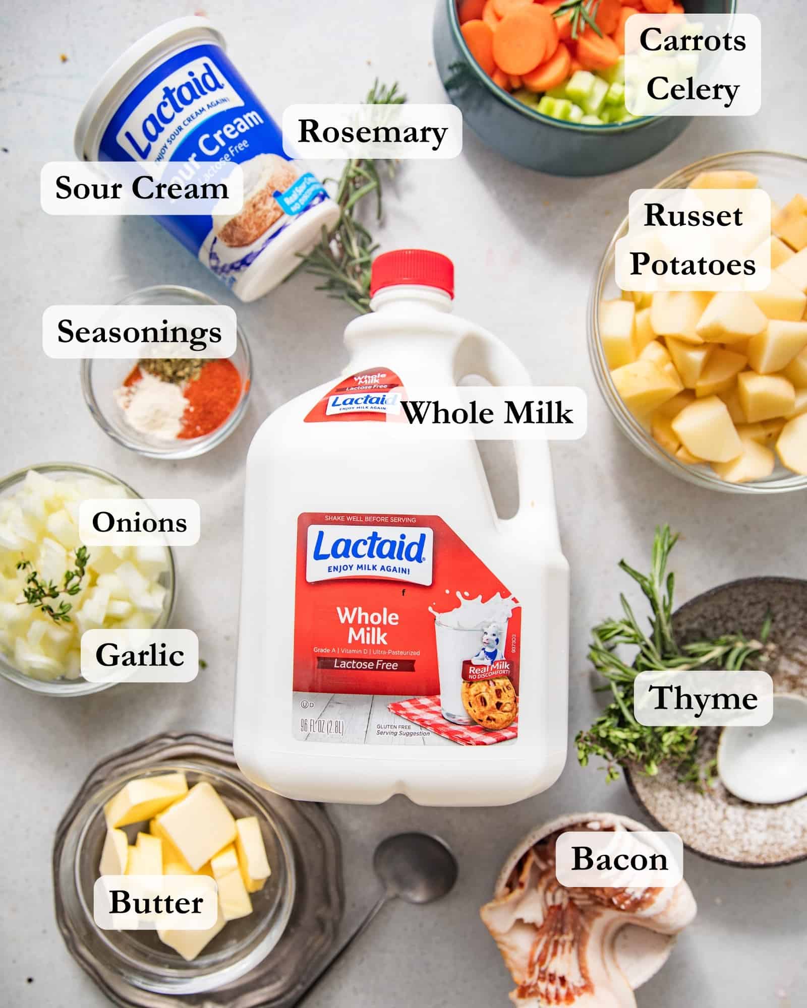 ingredients to make loaded baked potato soup on a white surface - russet potato, whole milk, bacon, butter, celery, carrots, thyme, rosemary, seasonings, onions, garlic, sour cream, and chicken bouillon paste.