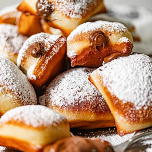 nutella beignets stacked on top of each other and covered in powdered sugar with nutella oozing from the center.
