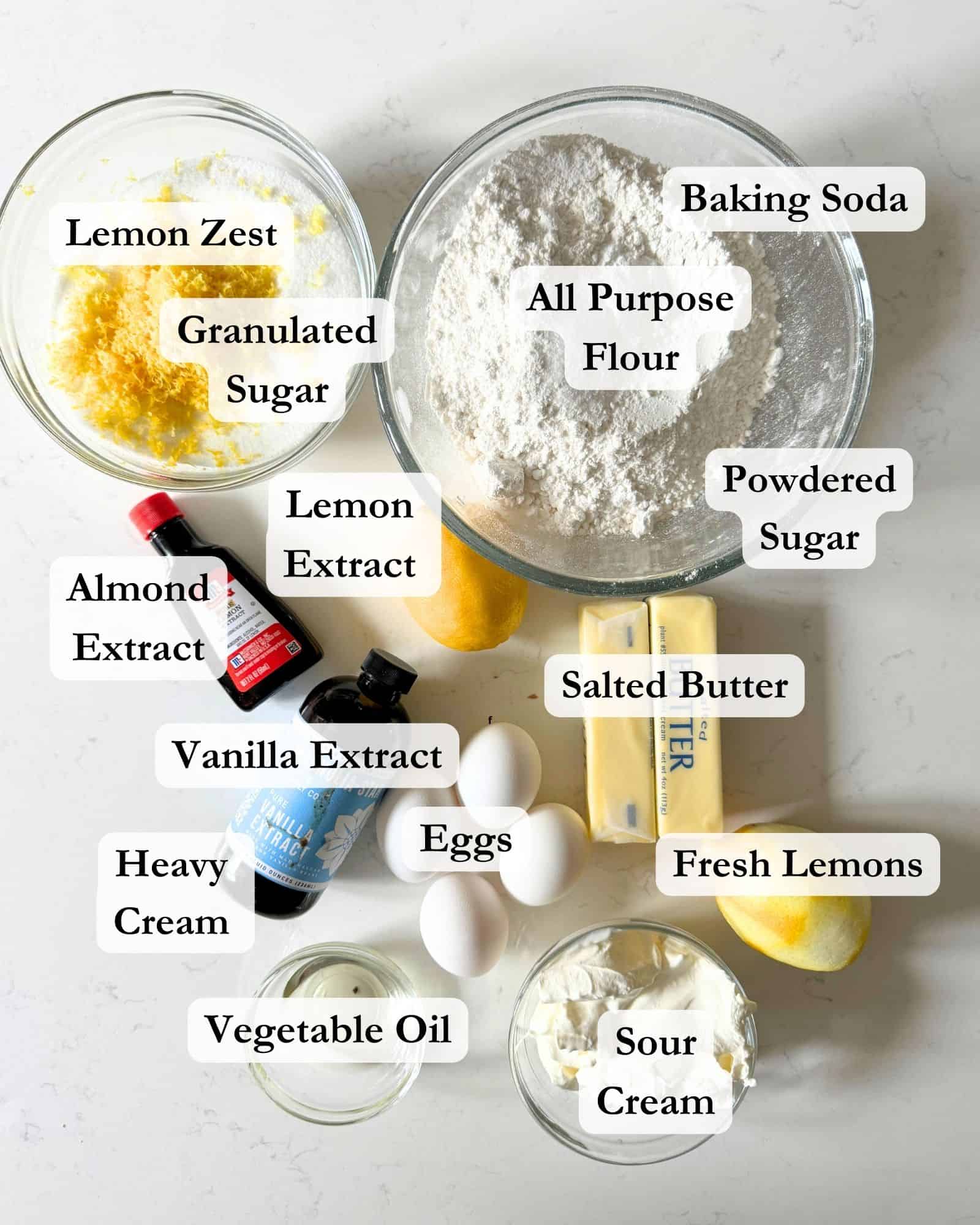 lemon pound cake and lemon glaze ingredients on a white surface - cake flour, butter, sugar, lemon zest, lemons, lemon extract, vanilla extract, almond extract, baking soda, kosher salt, sour cream, powdered sugar, heavy cream, and vegetable oil.