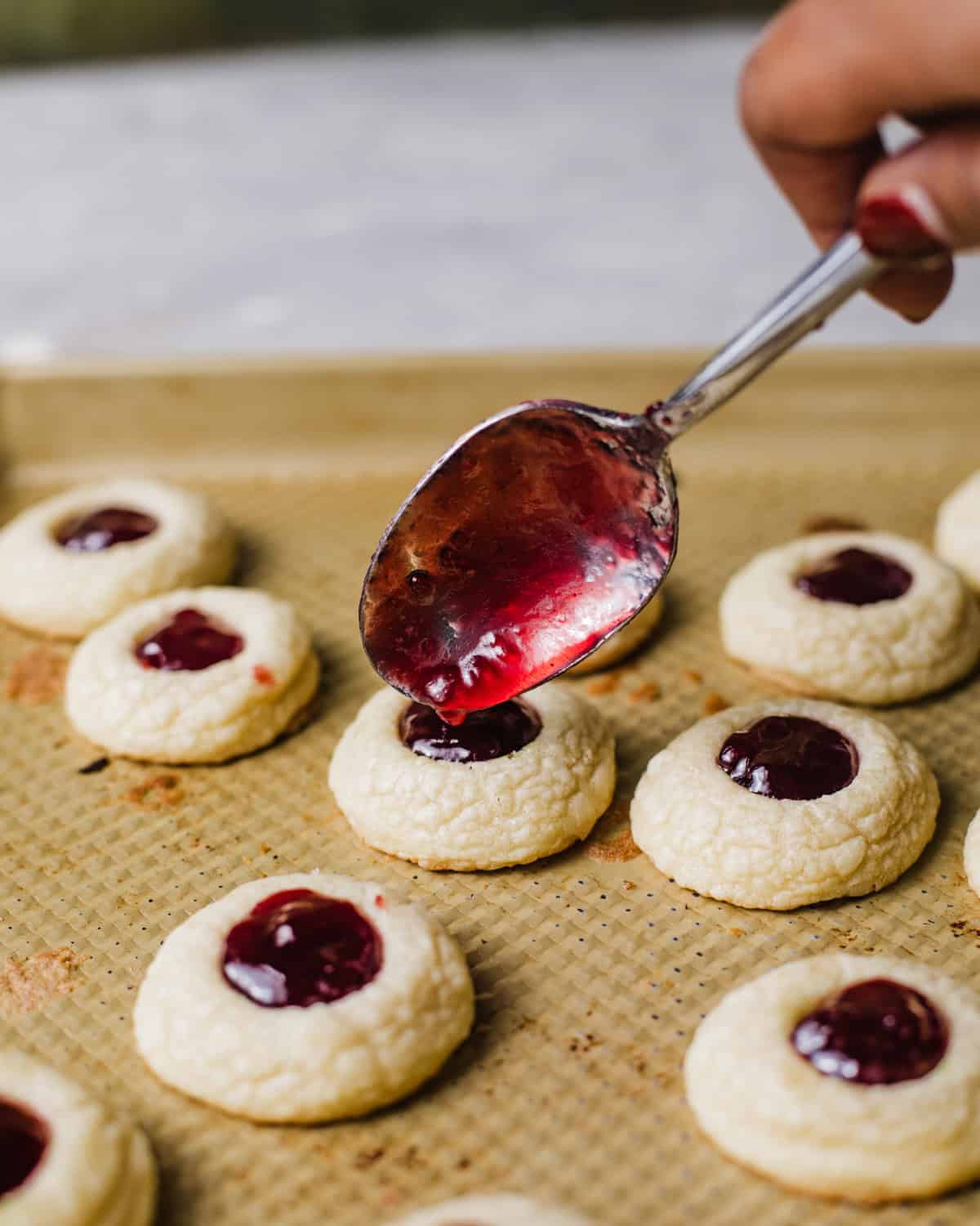 a spoon pouring raspberry jam into the center of the thumbprint cookies. 