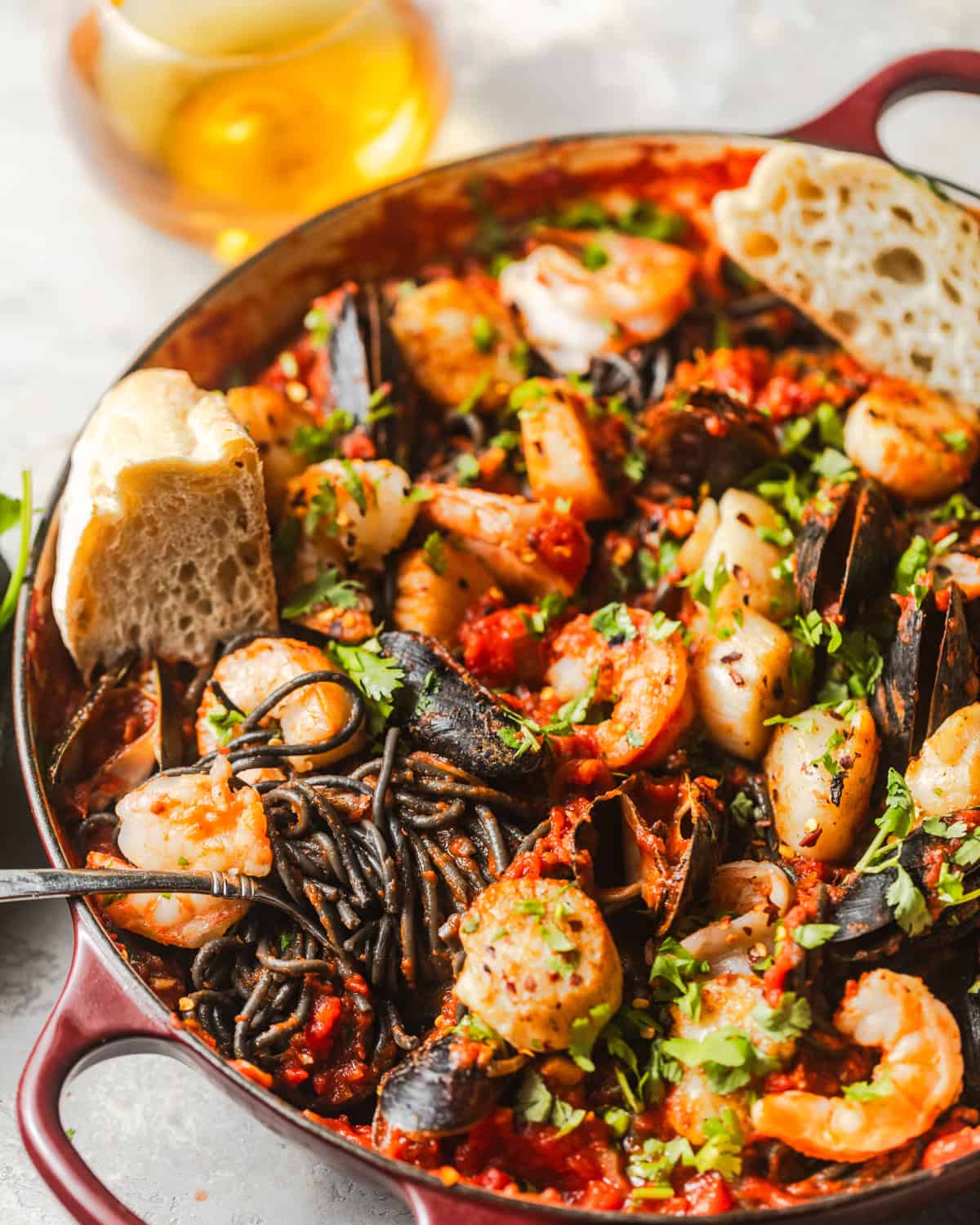 squid ink pasta in a skillet with shrimp, mussels, and scallops and a piece of crusty bread.