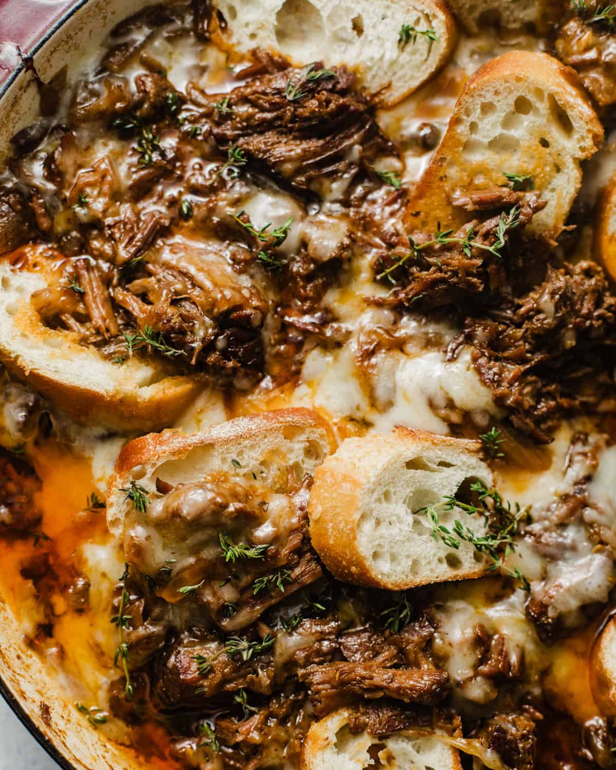 up close photo of french onion pot roast in a braising pan topped with gruyere cheese and sliced baguettes.