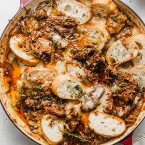 french onion pot roast in a skillet topped with gruyere cheese and sliced baguette bread.