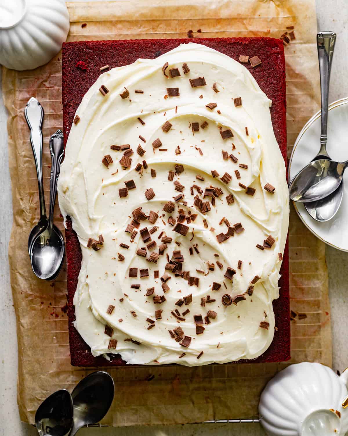 overhead photo of red velvet sheet cake on a piece of parchment paper topped with cream cheese frosting and chocolate shavings. 