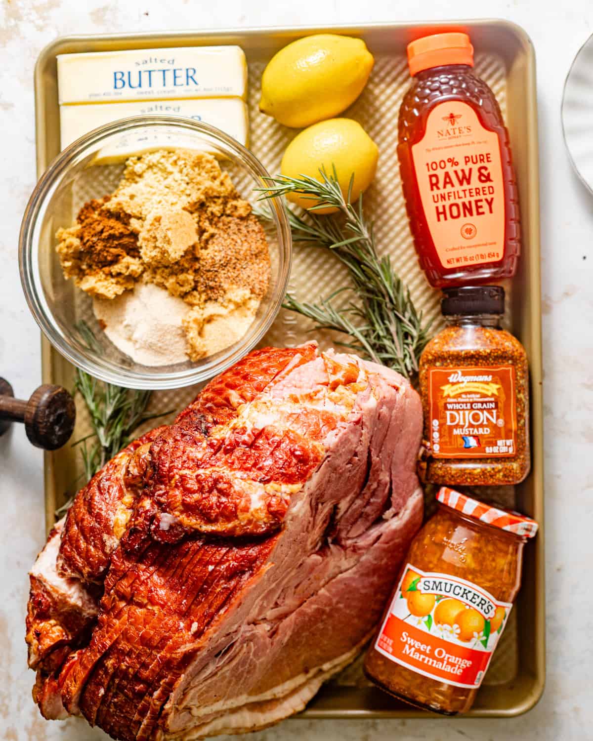 ingredients to make brown sugar mustard glazed ham on a baking sheet - bone in spiral ham, brown sugar, dijon mustard, butter, honey, orange marmalade, lemons, pineapple juice, and seasonings. 