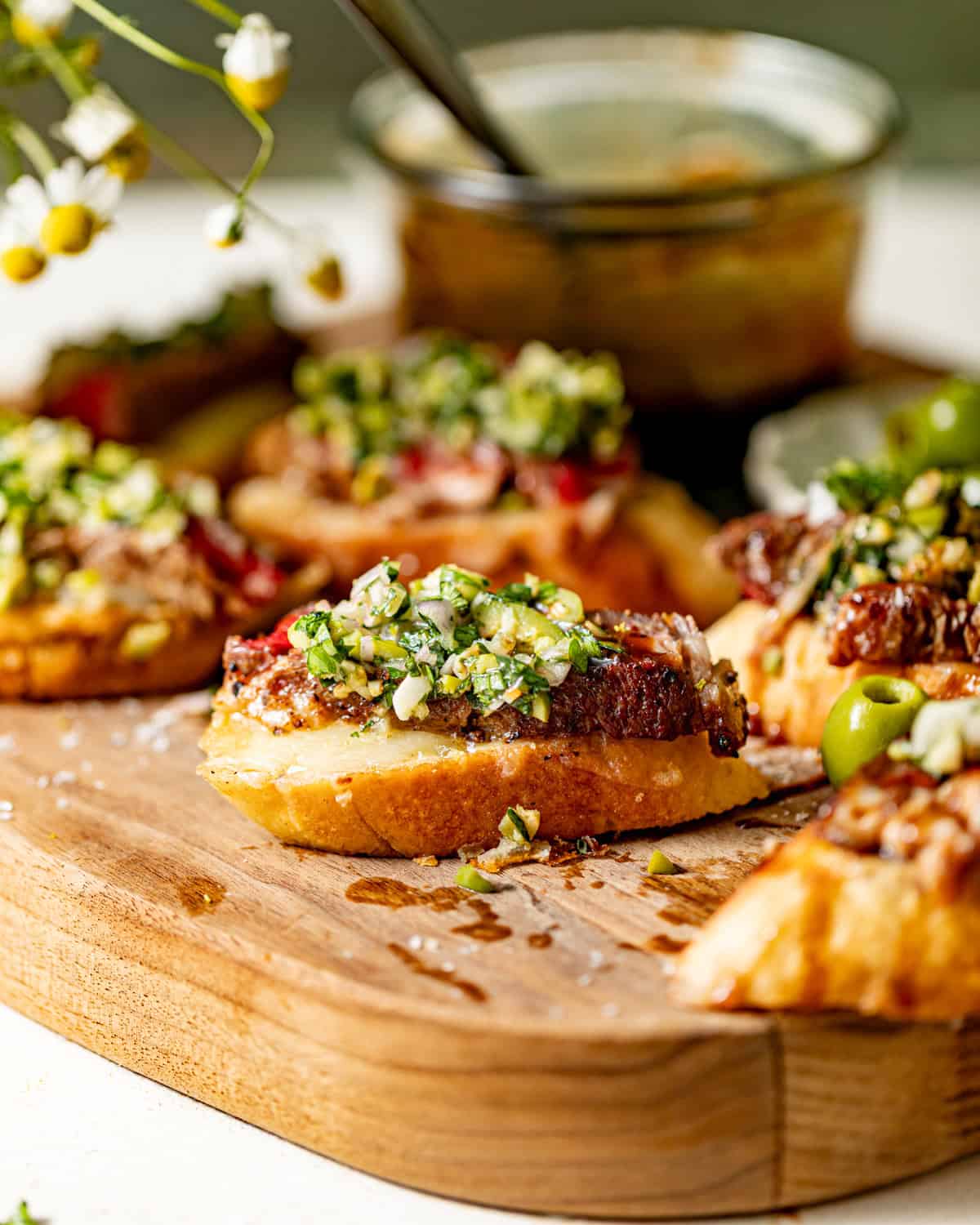 toasted crostini topped with mozzarella cheese, sliced steak, olive salsa, and balsamic glaze on a cutting board.