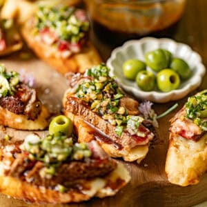 steak crostini on a cutting board in front of olives in a bowl.