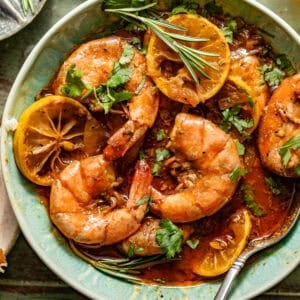 new orleans bbq shrimp in a bowl with a spoon garnished with fresh parsley and rosemary alongside a piece of crusty bread.
