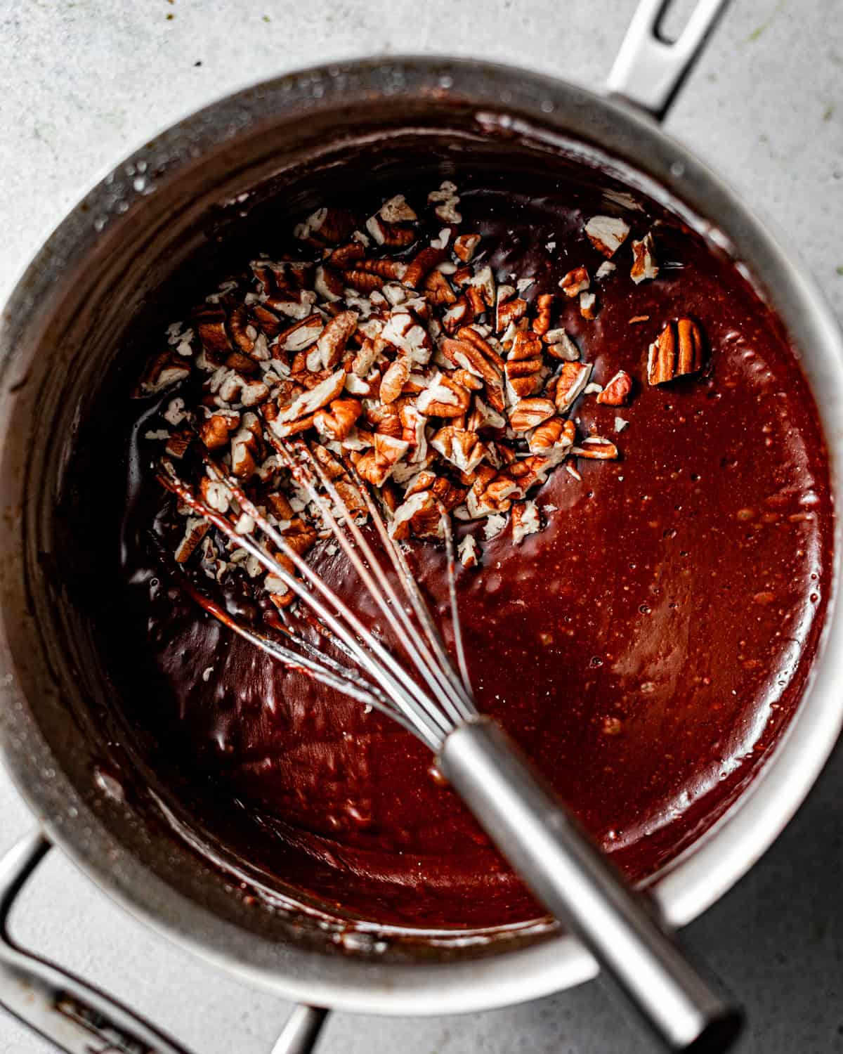 chocolate fudge frosting in a saucepan with chopped pecans and a whisk mixing everything together.