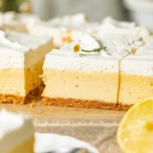 creamy lemon bars with graham cracker crust on a piece of parchment paper topped with edible flower and lemon whipped cream.