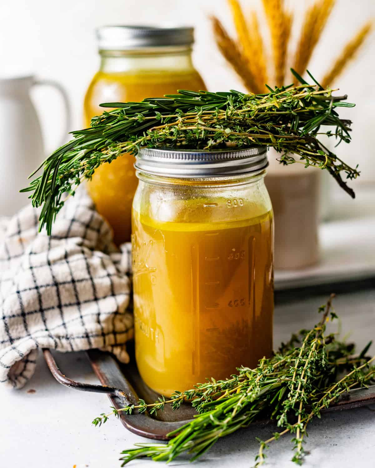 Mason jar of homemade chicken broth styled with fresh herbs, shredded chicken, and a second jar in the background.
