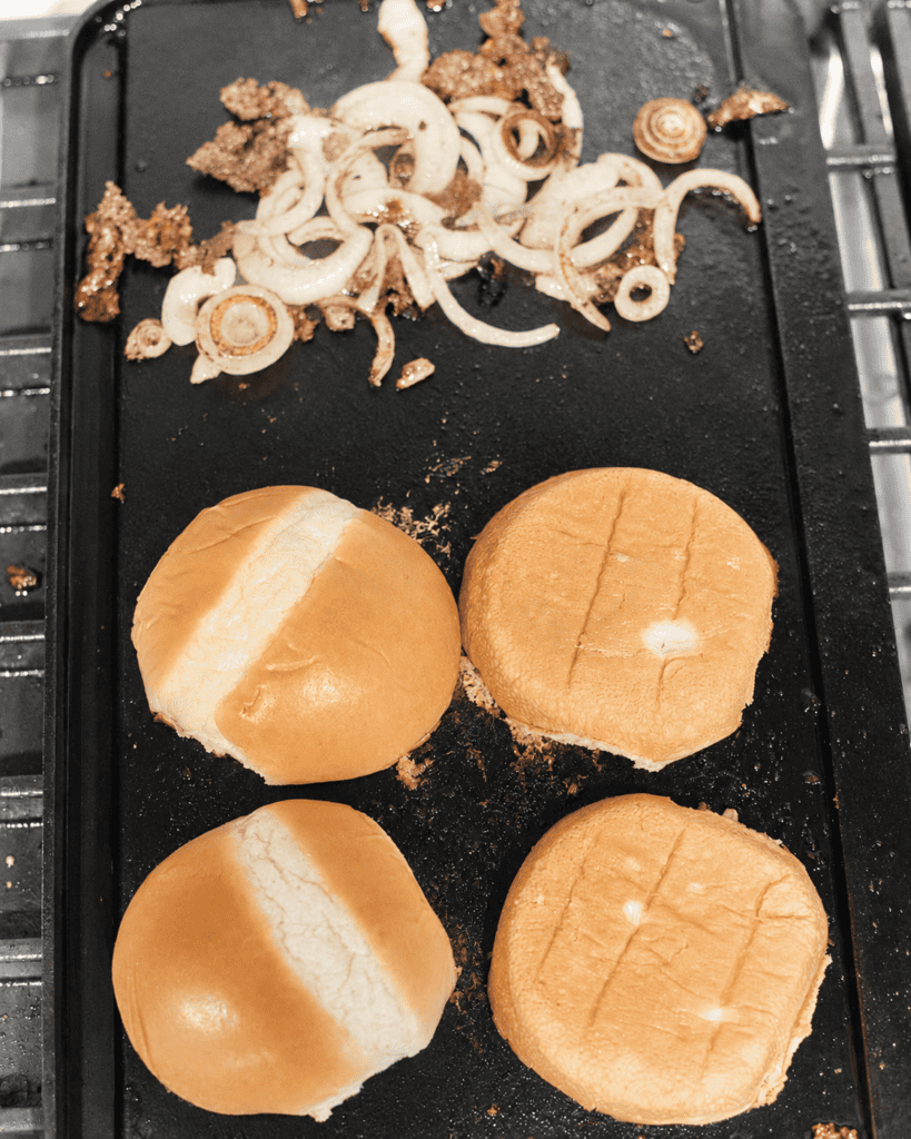toasted hamburger buns on a griddle with fried onions on the side.