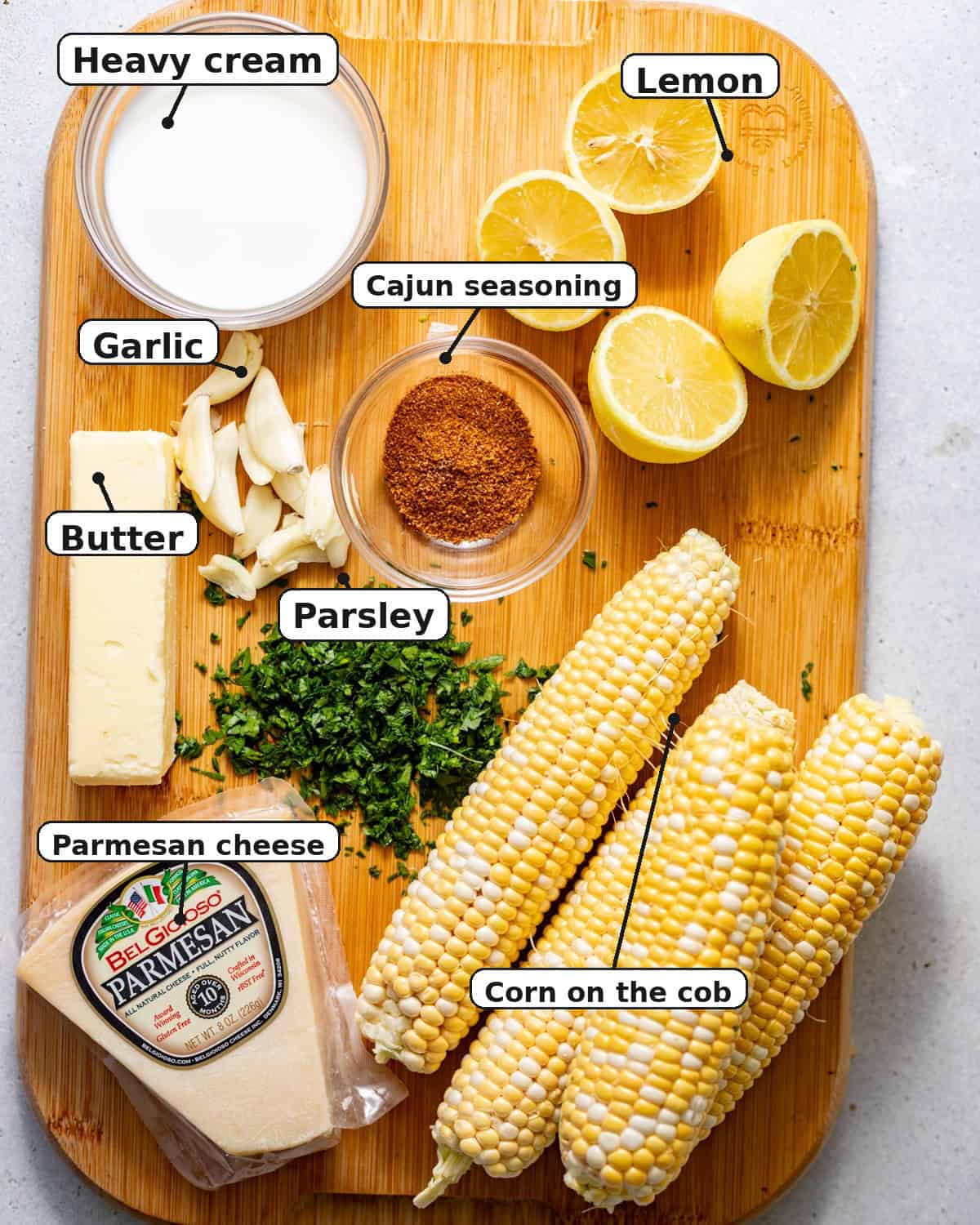 ingredients to make baked corn on the cob on a cutting board - fresh corn on the cob, seasonings, garlic, parsley, butter, heavy cream, lemons, and parmesan.