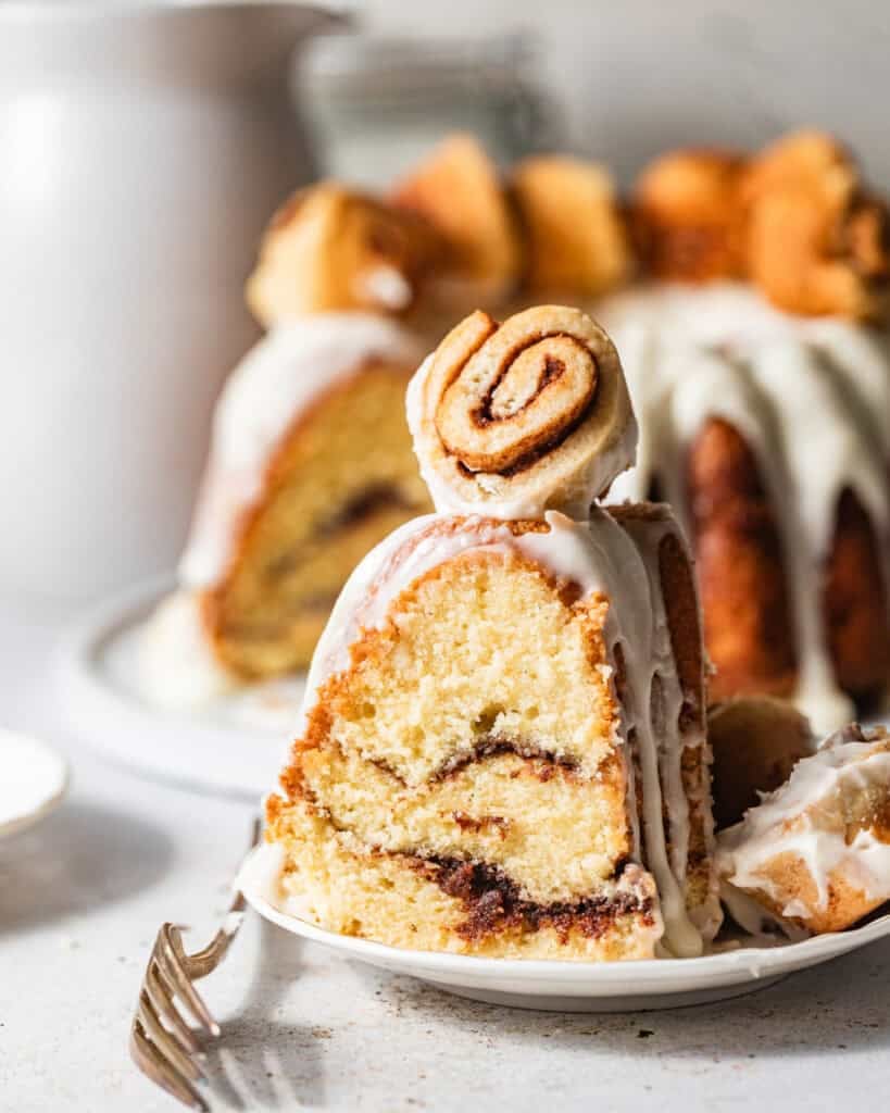 a slice of cinnamon roll pound cake on a cake plate topped with a mini cinnamon roll.