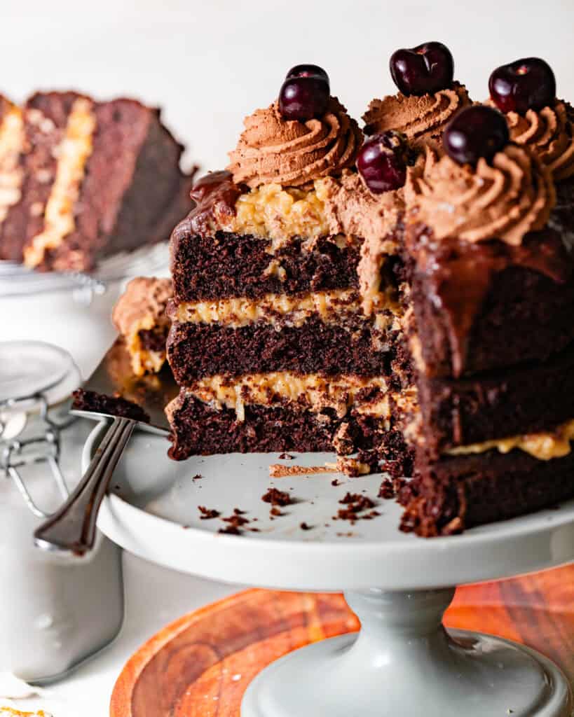 German chocolate cake with coconut pecan frosting on a cake stand with a cake server with a few slices taken out.