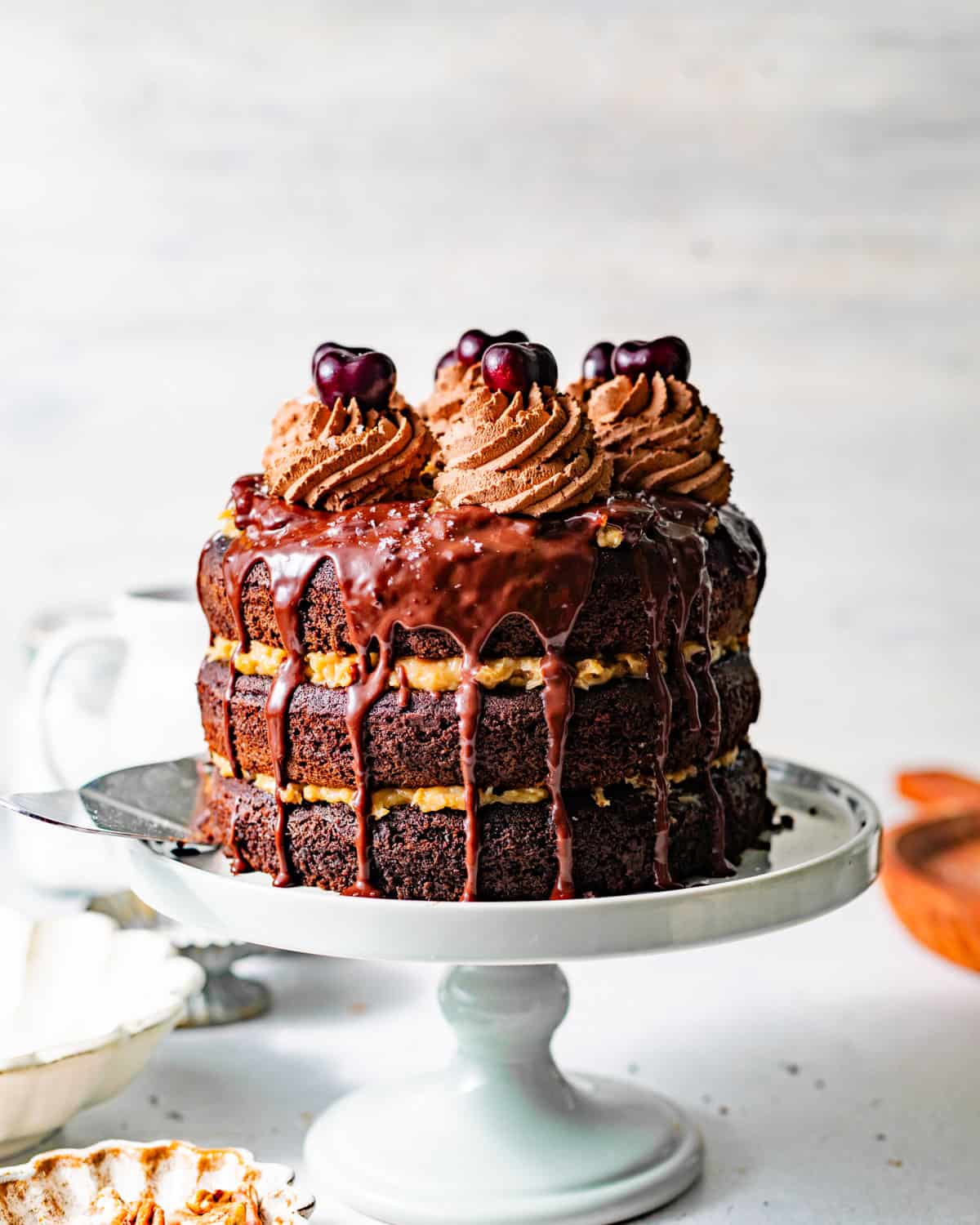 german chocolate cake on a cake stand with chocolate ganache and chocolate whipped cream and cherries on top.