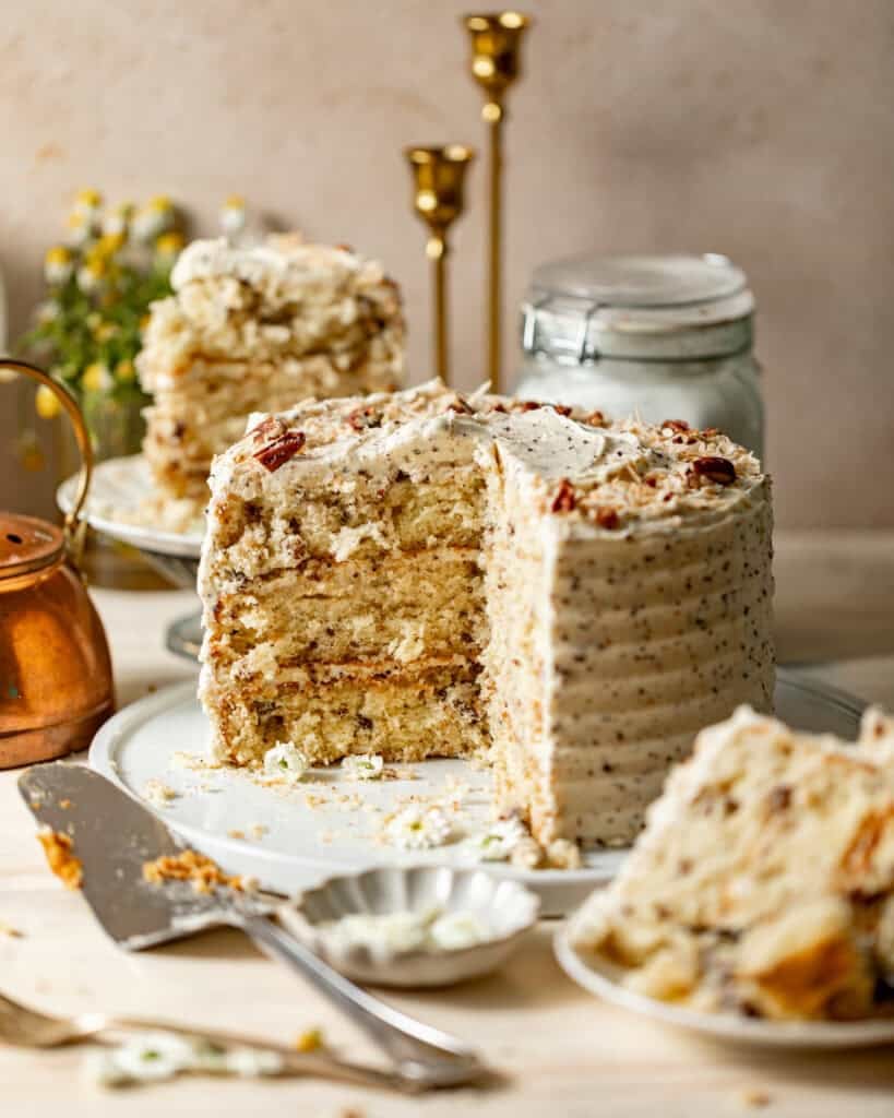 Italian cream layer cake with a slice removed, topped with chopped pecans and shredded coconut.