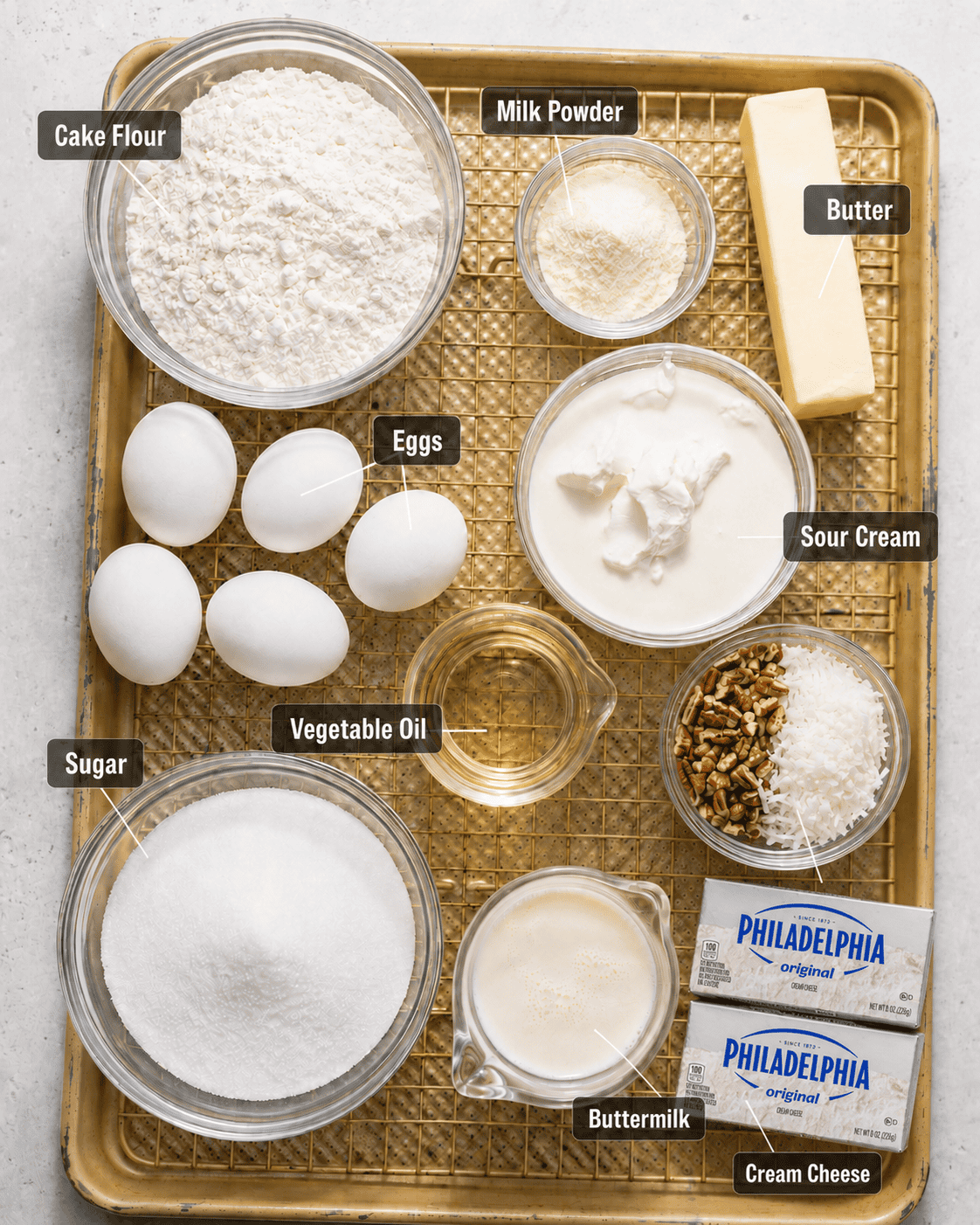 Italian cream cake main ingredients on a baking sheet in a small bowl including cake flour, sugar, milk powder, eggs, buttermilk, sour cream, pecans, coconut, vegetable oil, butter, and cream cheese.
