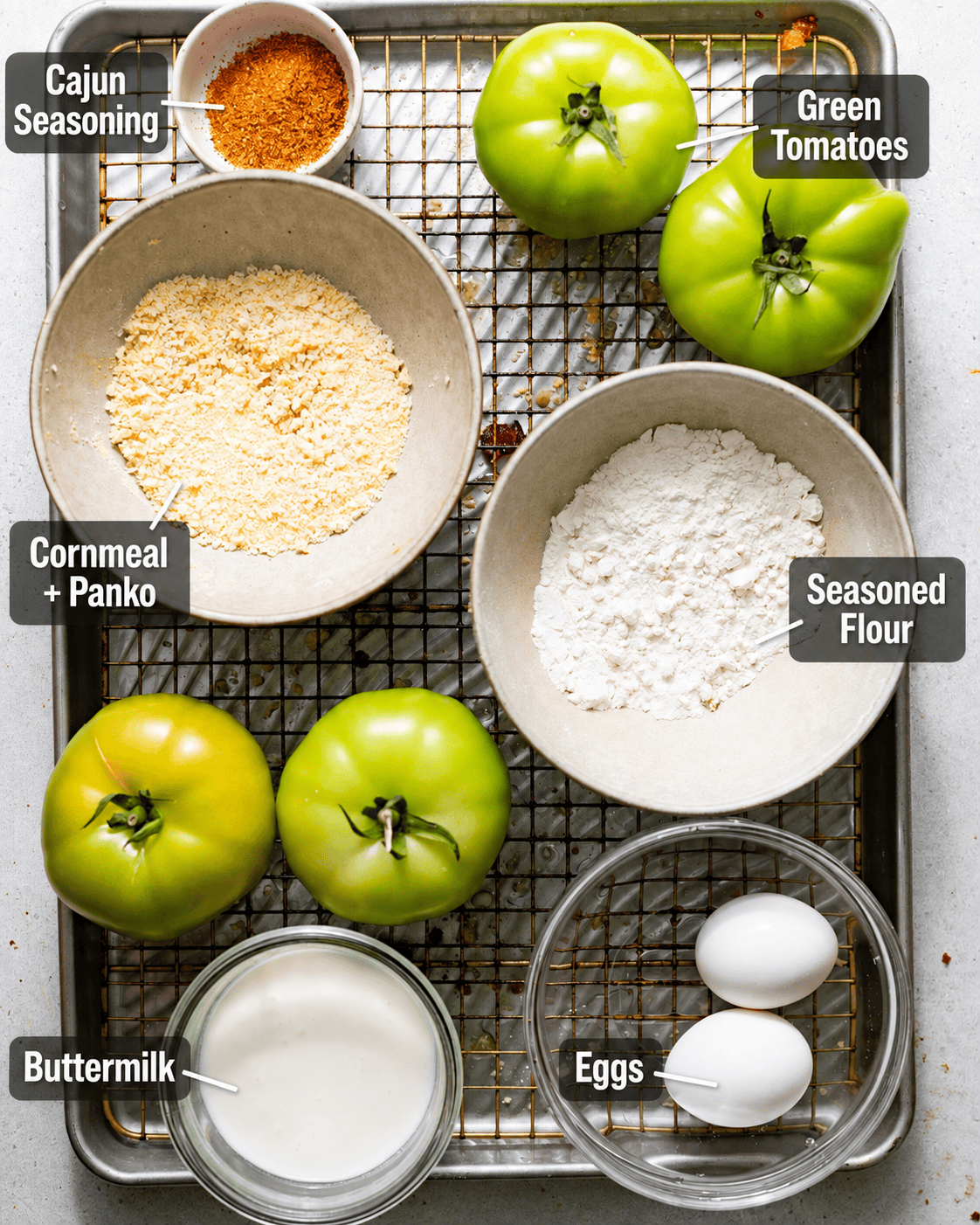 ingredients to make fried green tomatoes on a baking sheet - sliced green tomatoes, Cajun seasoning, flour, panko breadcrumbs, cornmeal, buttermilk, and eggs