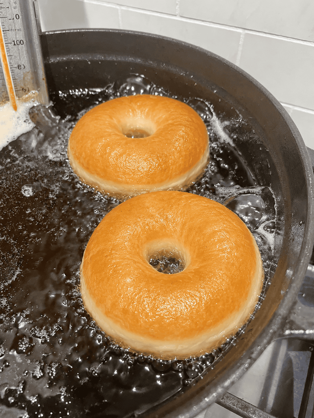 Two golden brown yeast donuts frying in bubbling oil with a candy thermometer in a dutch oven.