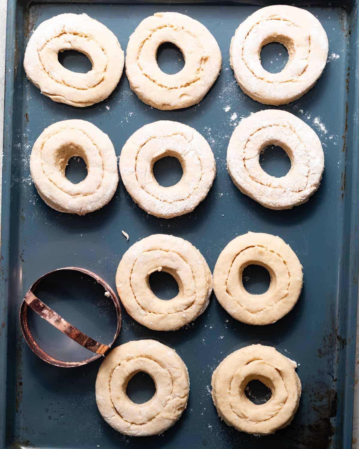 Unfried maple cake donuts with scored tops arranged on a baking sheet beside a round donut cutter.