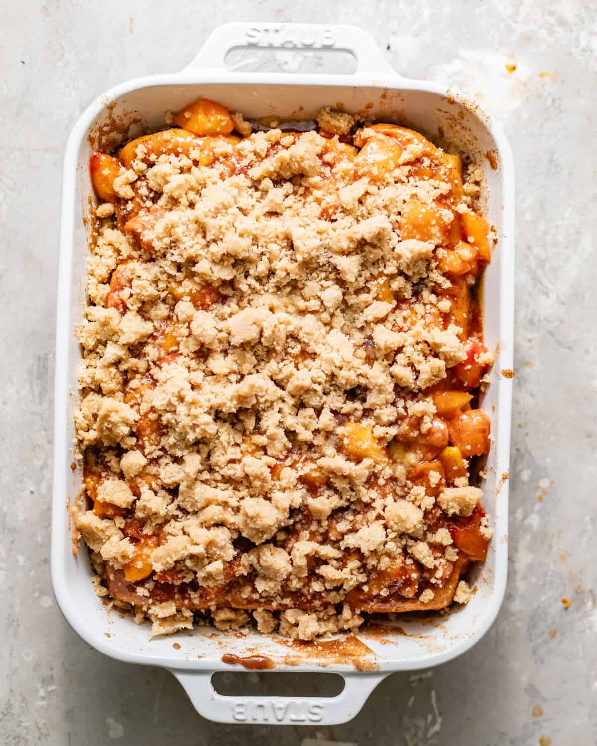 peach cobbler cinnamon roll bites assembled in a baking dish - hawaiian rolls covered with cinnamon roll filling and filled with peach cobbler and topped with streusel.