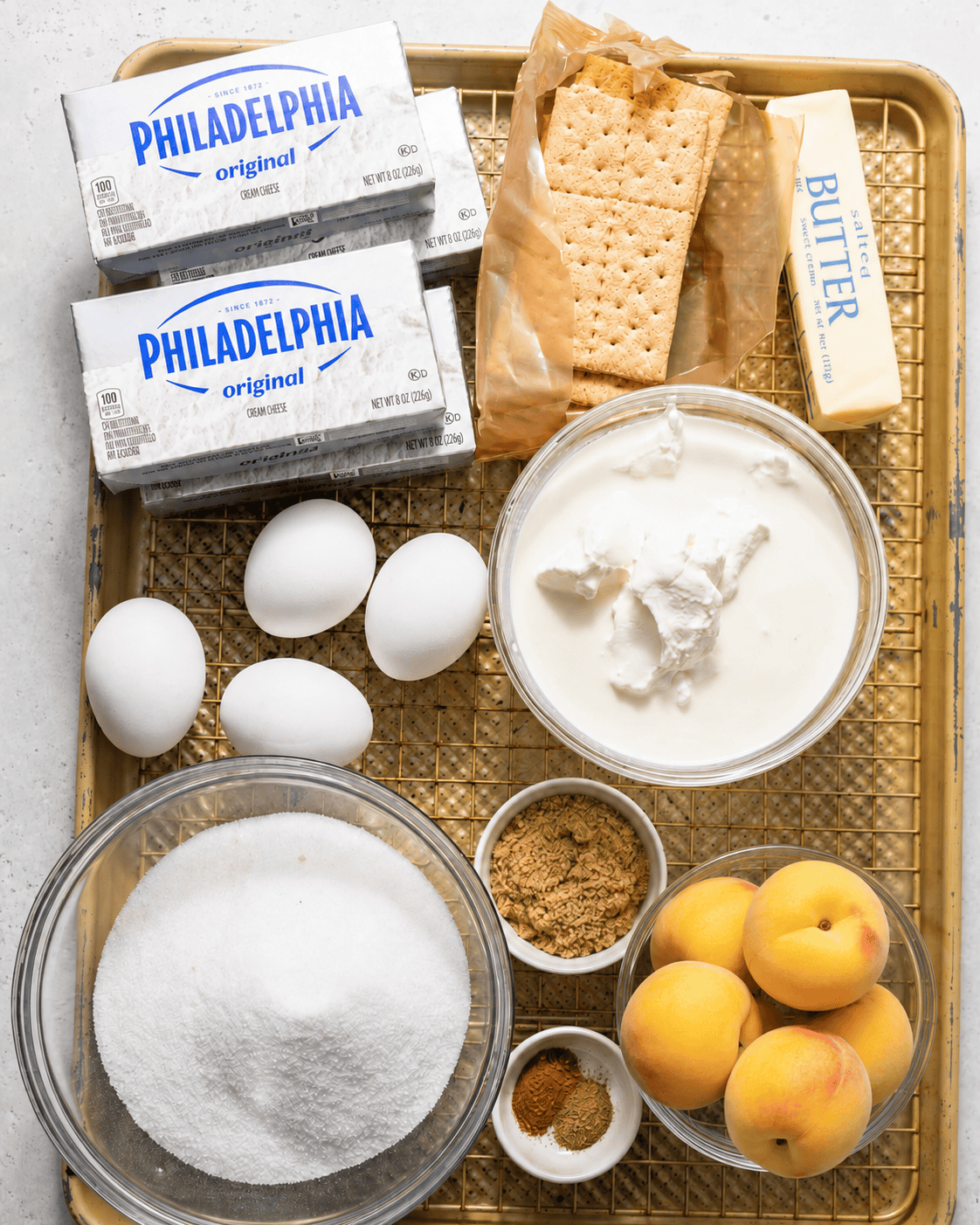 ingredients to make peach cobbler cheesecake on a baking sheet - fresh peaches, cream cheese, graham crackers, butter, heavy cream, sour cream, sug, brown sugar, eggs, and vanilla extract.