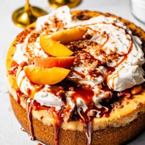 peach cobbler cheesecake on a white surface topped with peach cobbler, whipped cream, streusel, and salted caramel sauce.