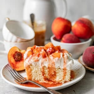a slice of peach cobbler tres leches on a plate topped with whipped cream, cooked peaches, and peach syrup.