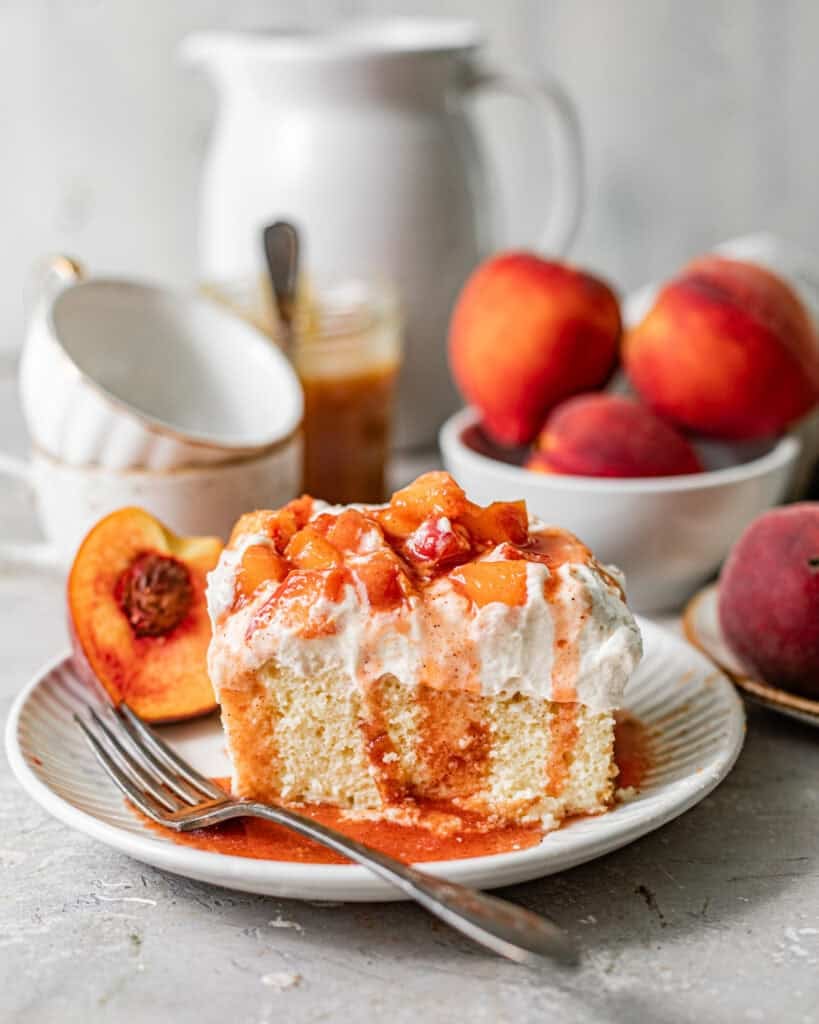 a slice of peach cobbler tres leches on a plate topped with whipped cream, cooked peaches, and peach syrup.