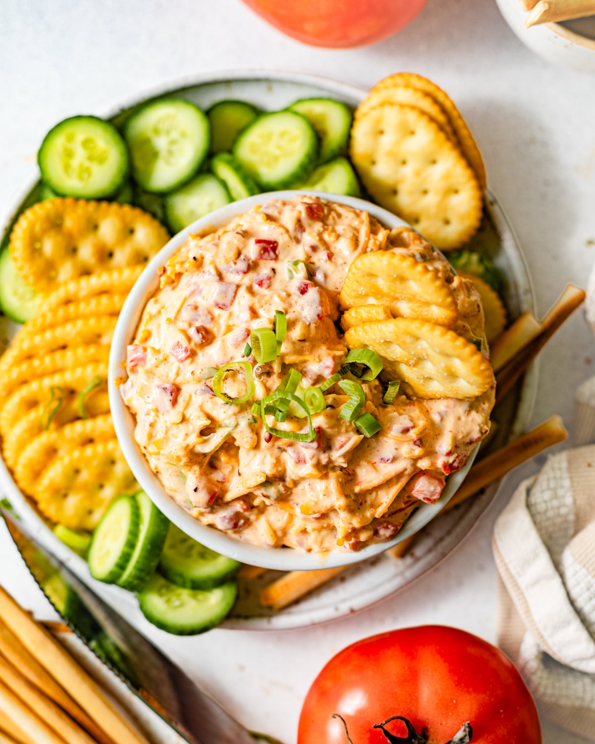 pimento cheese in a bowl next to sliced cucumbers, tomatoes, crackers, and breadsticks for dipping.