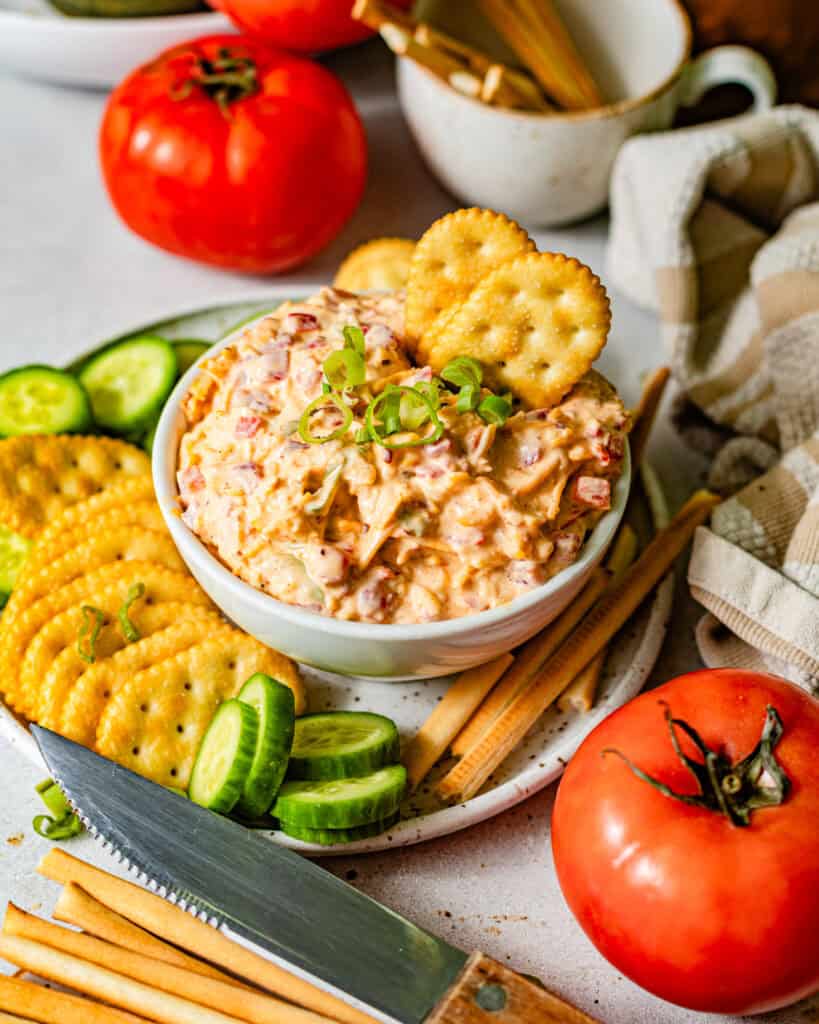 pimento cheese in a bowl surrounded by crackers, sliced cucumbers, and breadsticks.