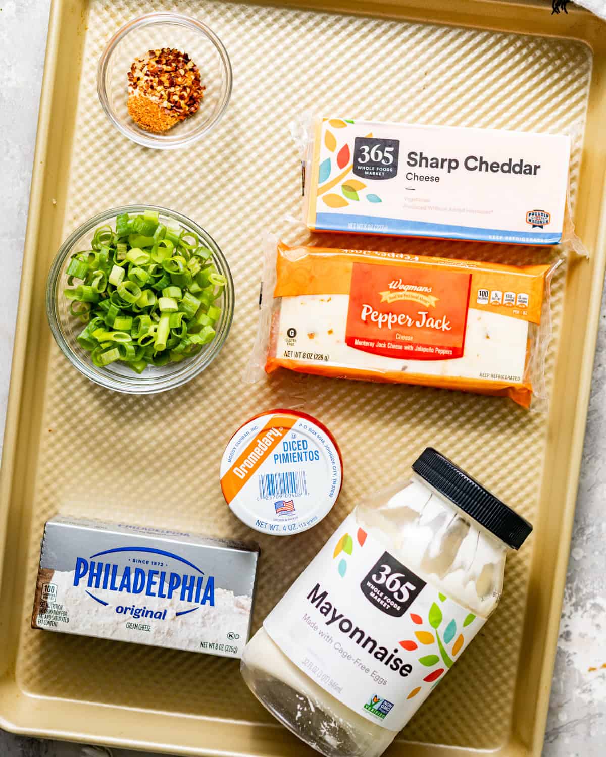 ingredients to make pimento cheese on a baking sheet - cheddar cheese, mayo, pepper jack cheese, scallions, diced pimentos, seasonings, and hot sauce.