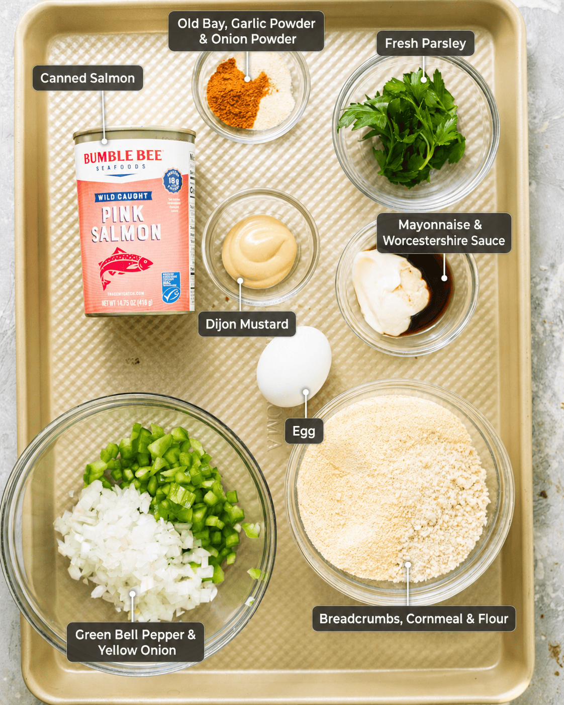 ingredients to make salmon croquettes on a baking sheet placed in small bowls - canned salmon, bell pepper, onion, parsley, seasonings, Dijon mustard, mayonnaise, Worcestershire sauce, egg, breadcrumbs, cornmeal, and flour with ingredient labels.