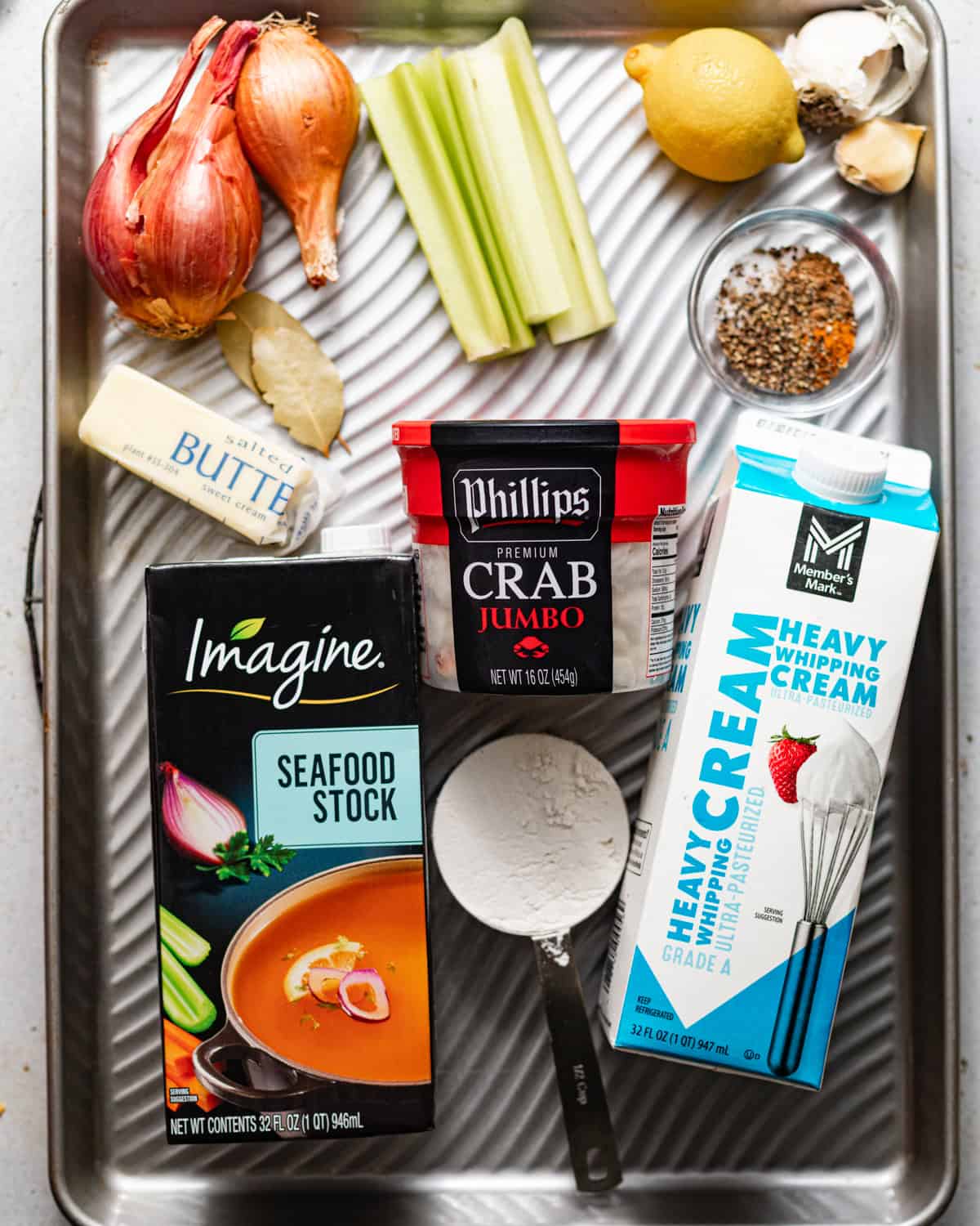 ingredients to make she crab soup on a baking sheet - jumbo lump crab meat, seafood stock, heavy cream, butter, flour, seasonings, shallots, celery, lemon, garlic, and bay leaves.