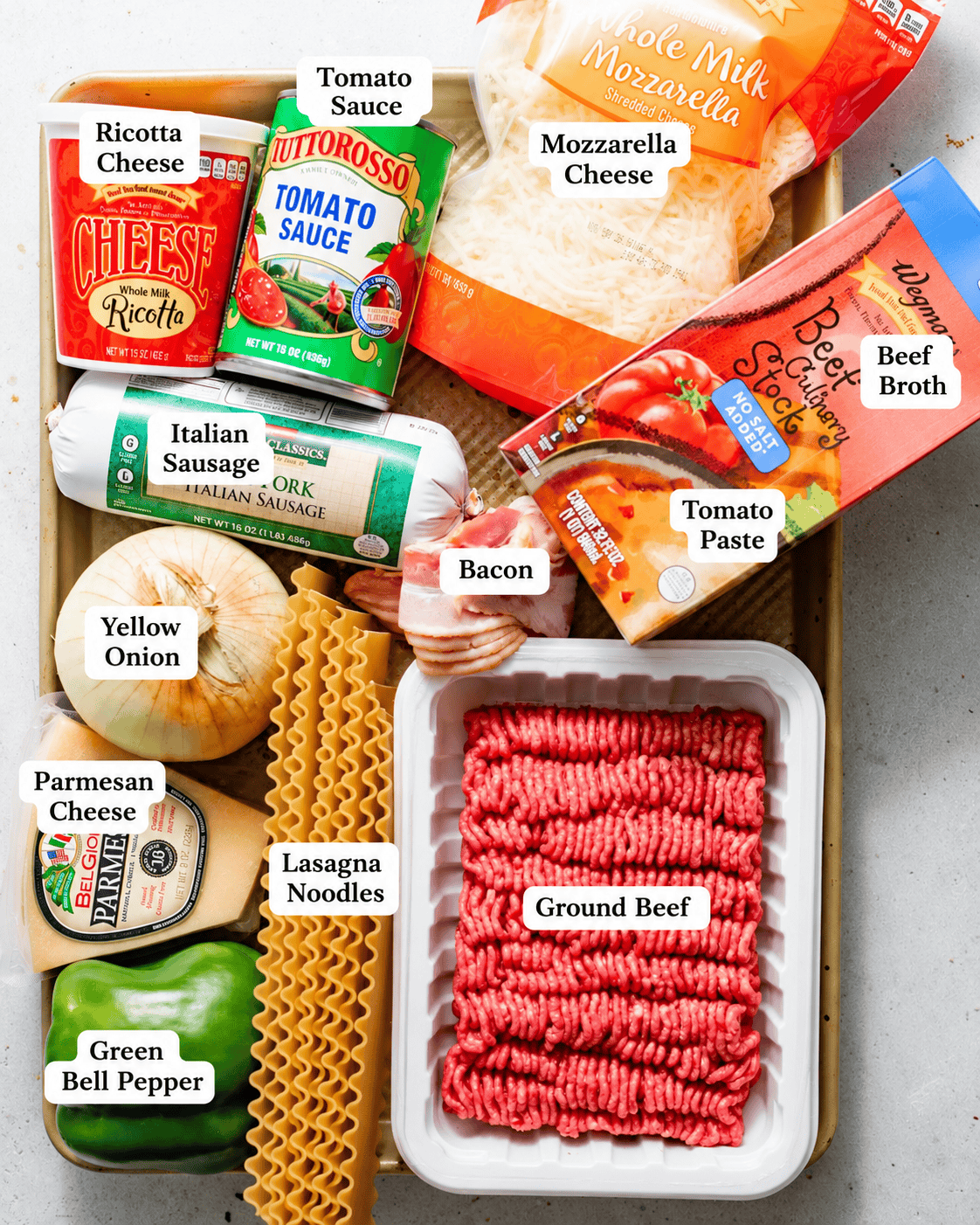 ingredients to make lasagna soup on a baking tray - ground beef, italian sausage, bacon, beef broth, tomato sauce, marinara, seasonings, green bell pepper, onion, garlic, parmesan, ricotta, heavy cream, and cheeses.