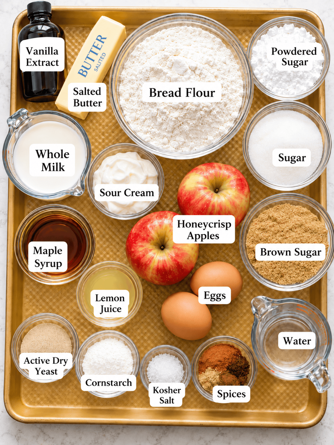 ingredients to make apple fritters on a baking sheet - bread flour, butter, sugar, brown sugar, sour cream, honeycrisp apples, vanilla extract, spices, maple syrup, active dry yeast, and spices.