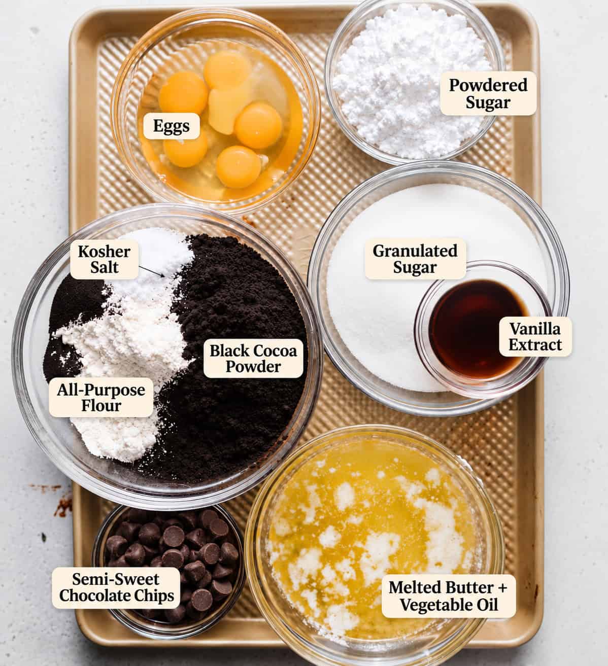 ingredients to make black cocoa brownies on a baking sheet - flour, eggs, sugar, powdered sugar, butter, vegetable oil, black cocoa powder, vanilla extract, and semi-sweet chocolate chips. 