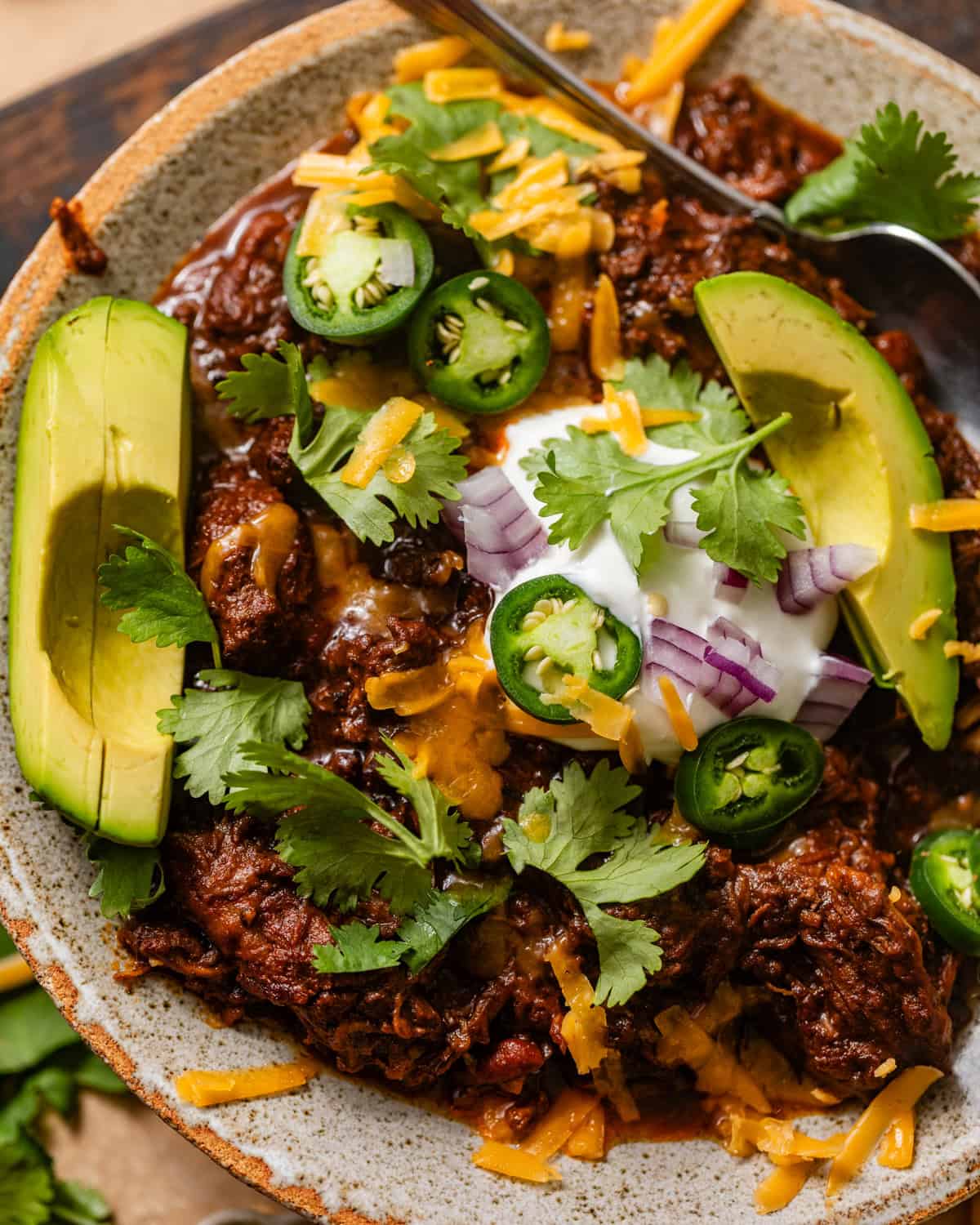 chuck roast chili in a bowl with various toppings and a spoon.