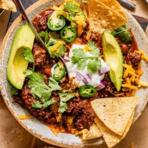 chuck roast chili in a bowl with tortilla chips and topped with sour cream, avocado, red onion, grated cheese, and cilantro.