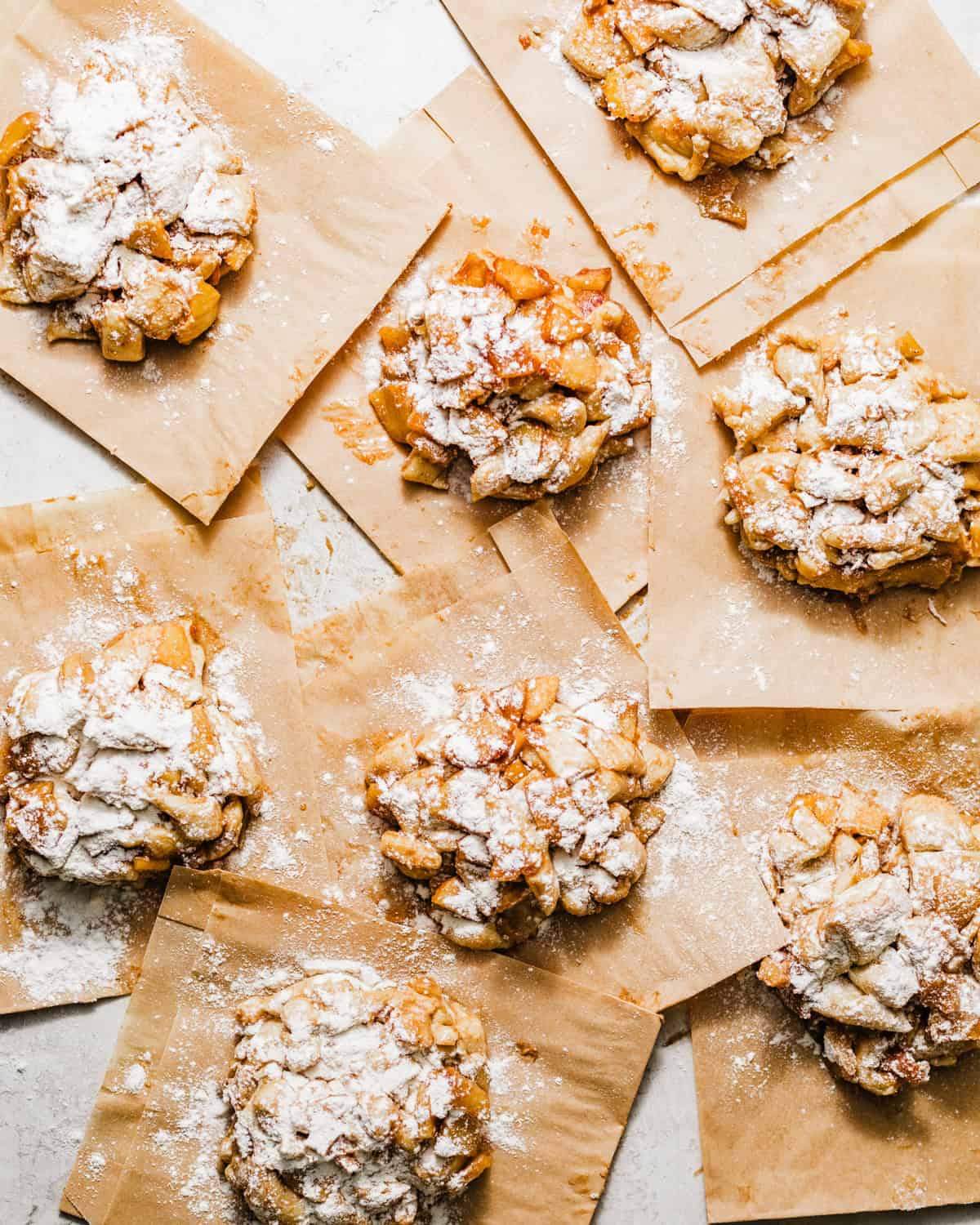 Shaped apple fritter donuts on parchment squares, filled with cinnamon-spiced apples and dusted with flour before frying.