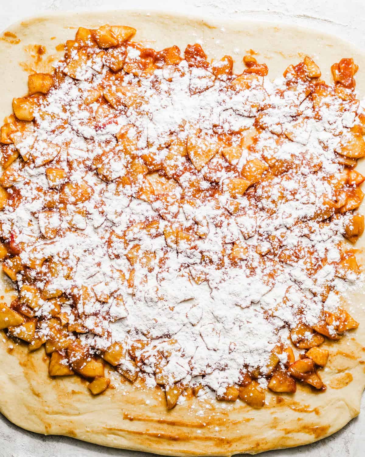 apple fritter dough on a flat surface with apple filling on top sprinkled with flour and cinnamon.