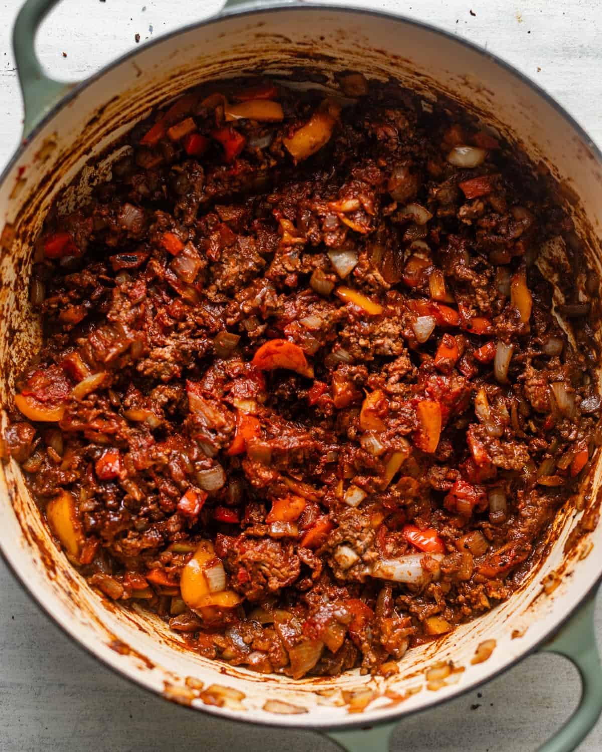 Browned ground beef, diced onions, bell peppers, and tomato paste mixed with chili seasonings in a Dutch oven before adding the broth and beans.
