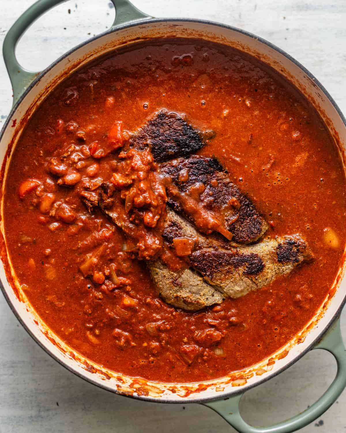 Seared chunks of beef chuck roast simmering in a tomato chili sauce with ground beef and seasoned beans in a Dutch oven.