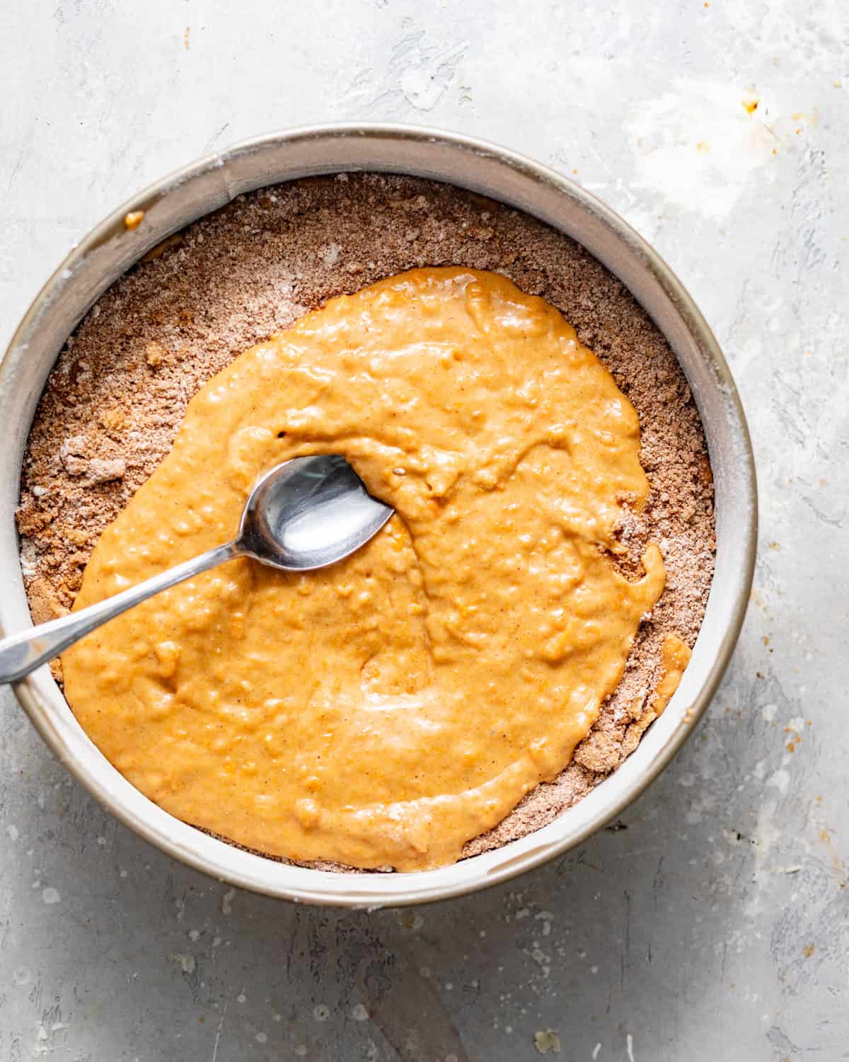 half of the cake batter added to a springform pan with cinnamon sugar filling sprinkled on top and a spoon adding the remaining batter to cover the filling.