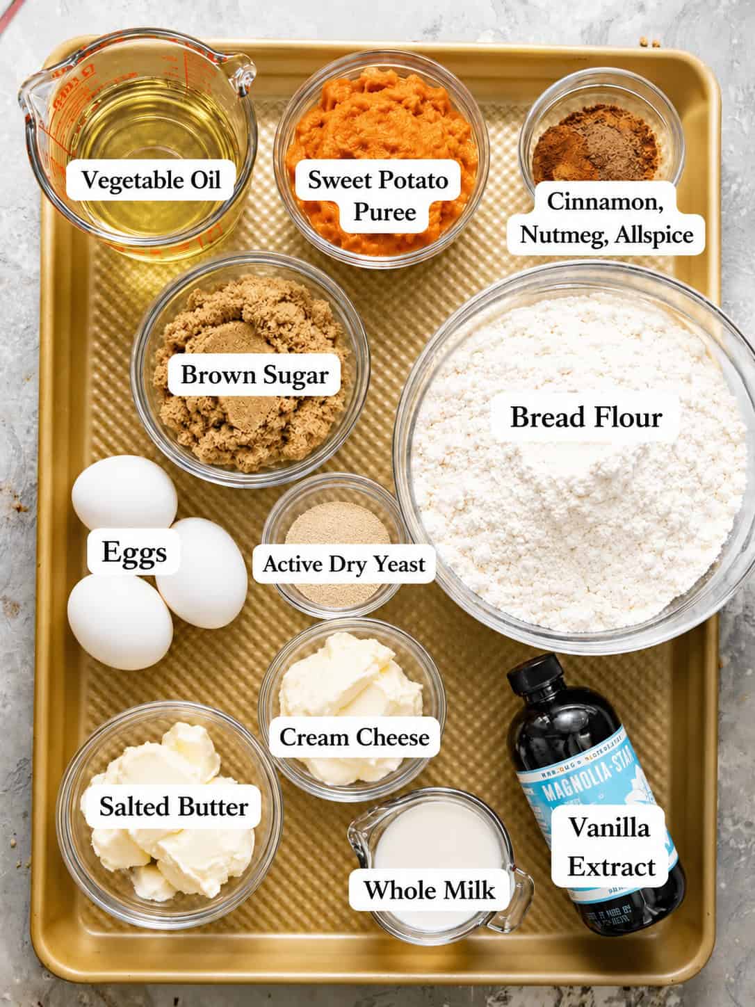 Labeled ingredient photo for sweet potato cinnamon rolls showing sweet potato puree, bread flour, brown sugar, vegetable oil, eggs, butter, maple syrup, sour cream, vanilla extract, baking powder, and cinnamon-spice mixture arranged in bowls on a gold sheet pan.